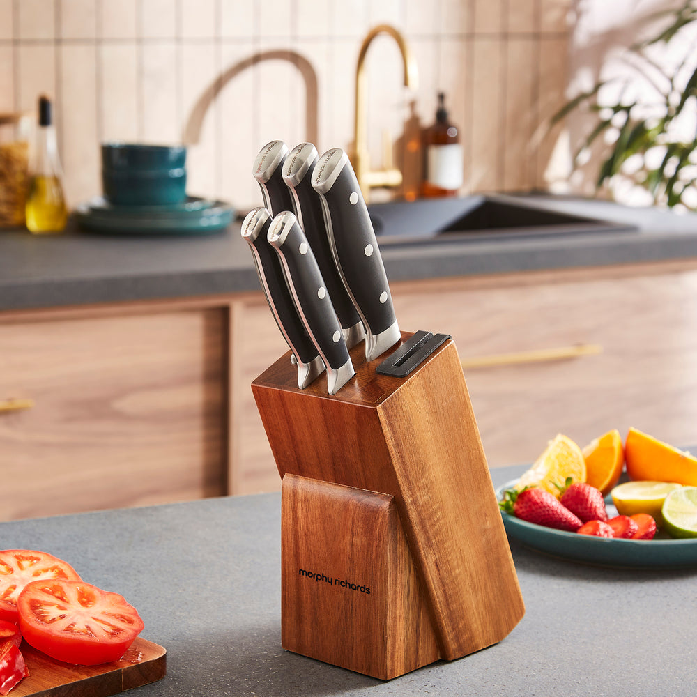 Set of knives in a wooden block on a kitchen counter with fruits and vegetables.