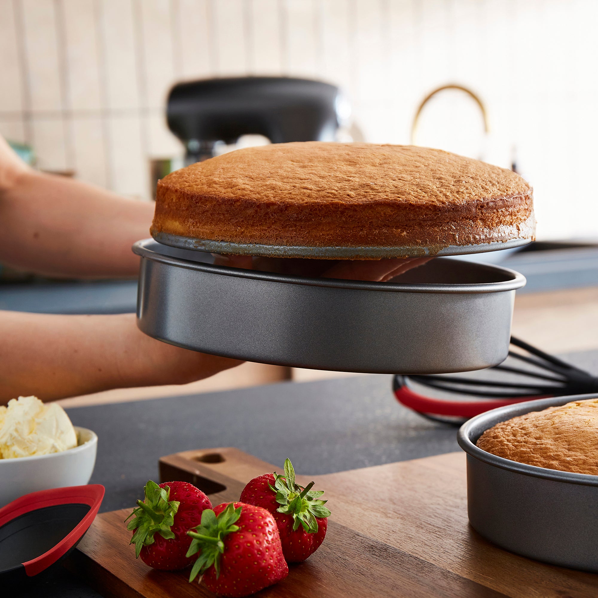 Baked cake in a round metal baking pan on a kitchen counter with strawberries and cream.