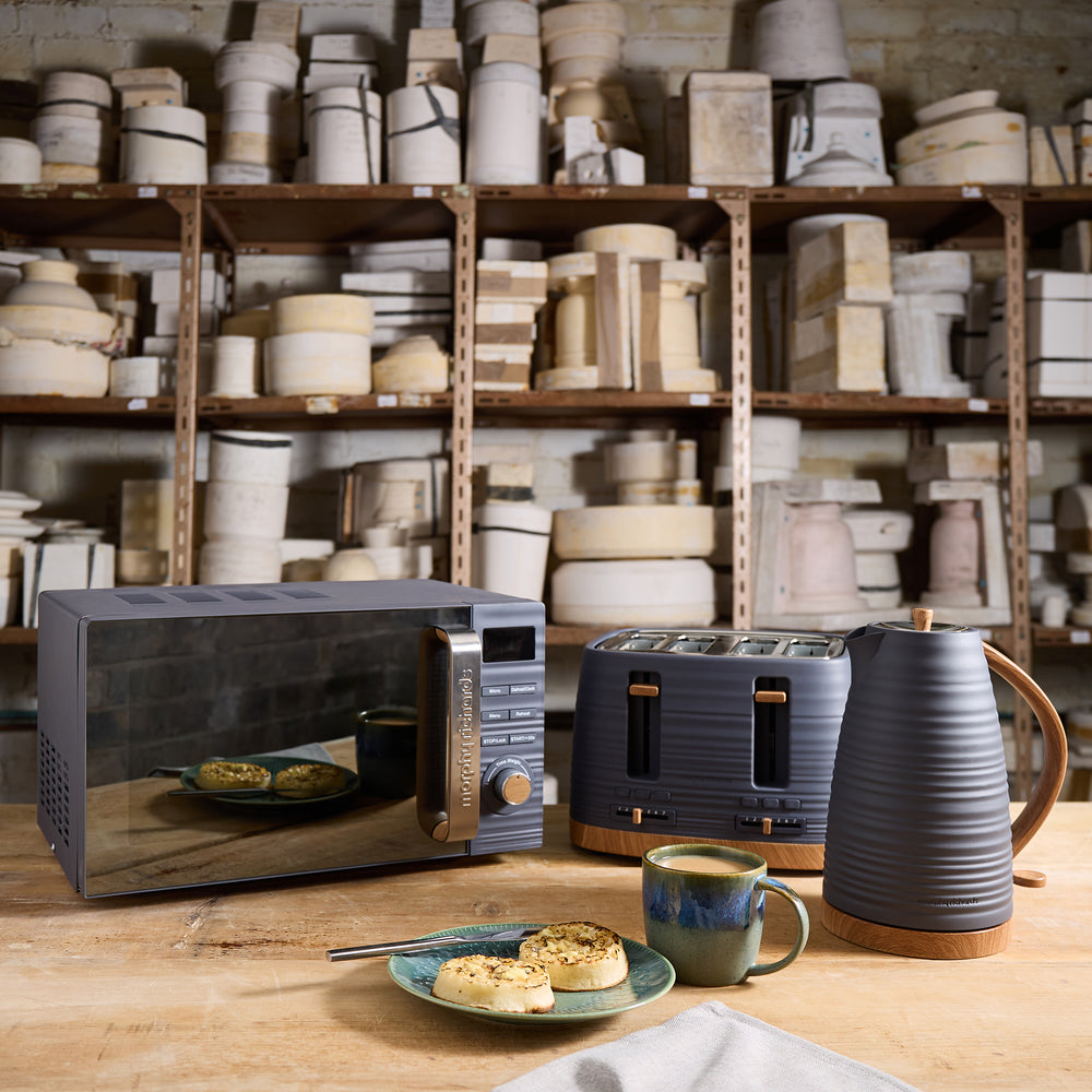 Set of kitchen appliances including a microwave, toaster, and kettle on a wooden table with shelves of ceramic items in the background.